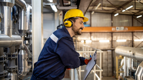 Mann in Arbeitskleidung und mit gelbem Schutzhelm lehnt an Reling und schaut in eine Industriehalle